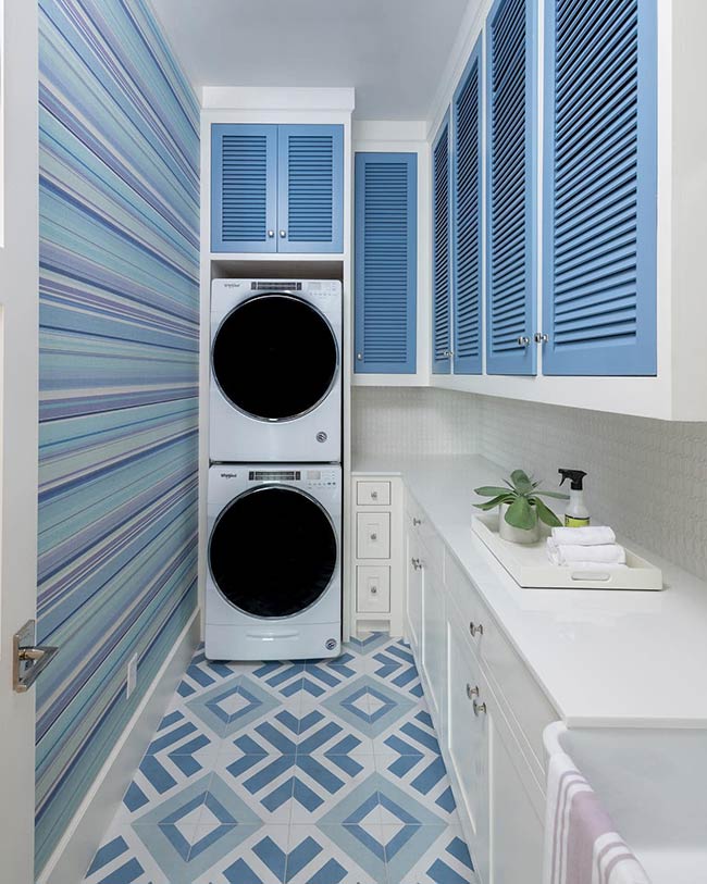 Striped wallpaper and geometric floor tiles create a sense of height and movement. White marble countertops and blue shutter cabinets complement the lower shaker-style cabinets. Even in a compact space, vertical and horizontal contrasts make the laundry room visually interesting.