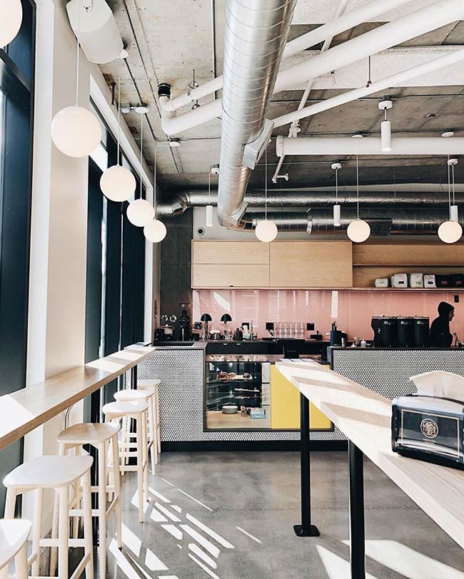 Glossy pink tiles add a sweet and soft touch to the back wall. The big silver pipes and hanging globe lights on the concrete ceiling keep the coffee shop interior looking modern and cool.