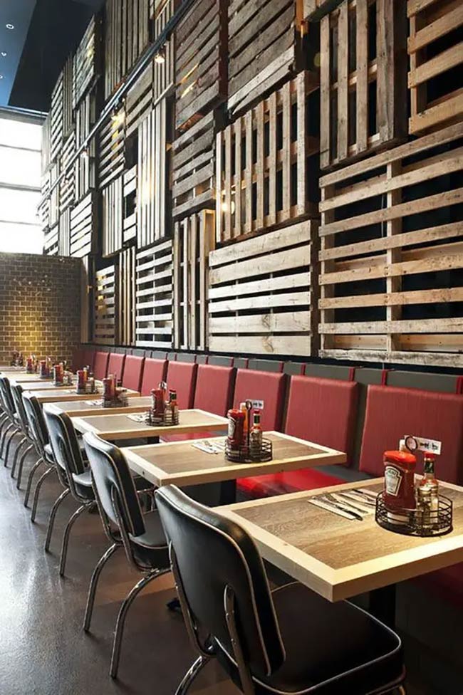 The back wall uses salvaged wooden pallets as a striking, textured focal point. Deep red booth seating runs along the wall. Classic black and white chairs blend the industrial wood with a retro flair.
