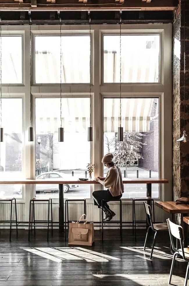 Tall, multi-paned windows illuminate the long window bar, which is perfect for solo work. Sleek, minimalist pendant lights hang low over the counter seats. The dark wood floor and exposed brick on the side walls anchor this industrial look.