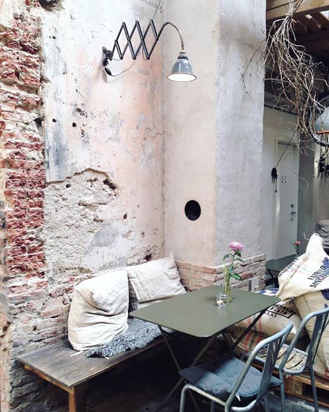 This is a tiny, incredibly charming corner defined by its distressed walls. Exposed brick blends with peeling plaster for a raw, aged texture. An adjustable industrial lamp extends from the wall, illuminating the simple outdoor furniture setup.