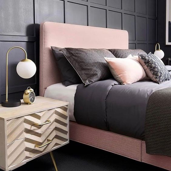 That wooden nightstand with the zig-zag carving is a beautiful piece. It sits nicely next to the soft pink headboard and the dark wall. The small gold lamp adds just the right amount of shine to this pink and black bedroom.