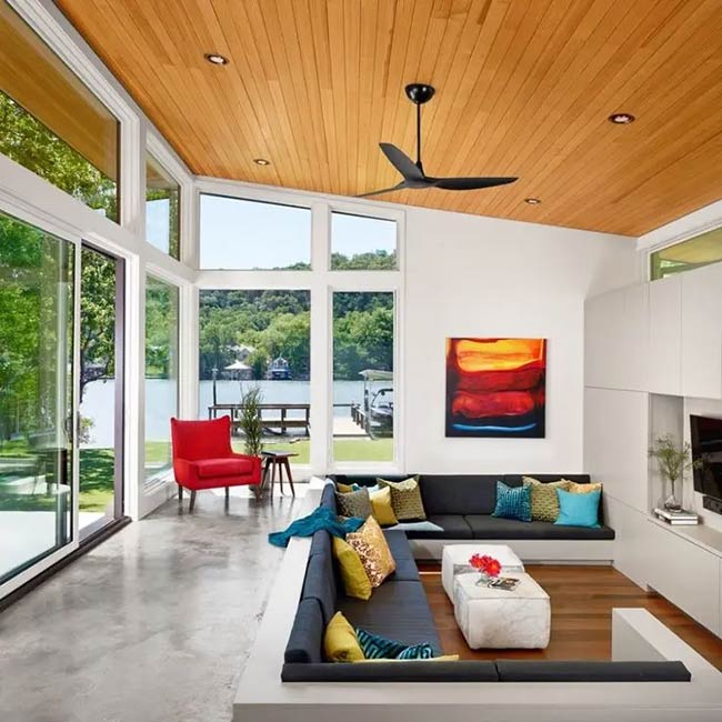 This conversation pit has a beautiful view of the lake. The dark sofa has bright yellow and blue pillows. The ceiling is made of wood.