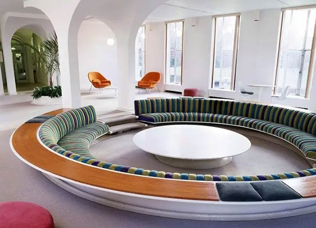 What a neat, circular conversation pit! The sofa has colorful stripes and a nice wood trim. It's in a big, bright white room with arches.