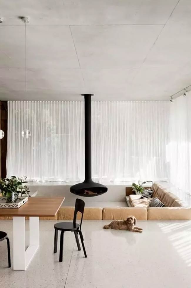 A little dog is lying on the floor right next to this sunken sofa. The black hanging fireplace is very modern. This whole room is very simple and white.