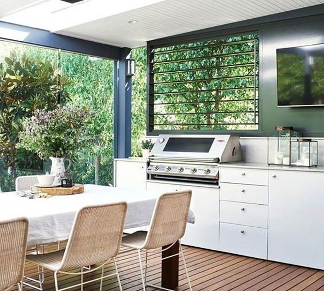 White cabinets give this covered deck a very clean and bright look. The stainless steel grill fits perfectly under the black shuttered window to let smoke out. Having a dining table right next to the cooking station makes serving meals easy.