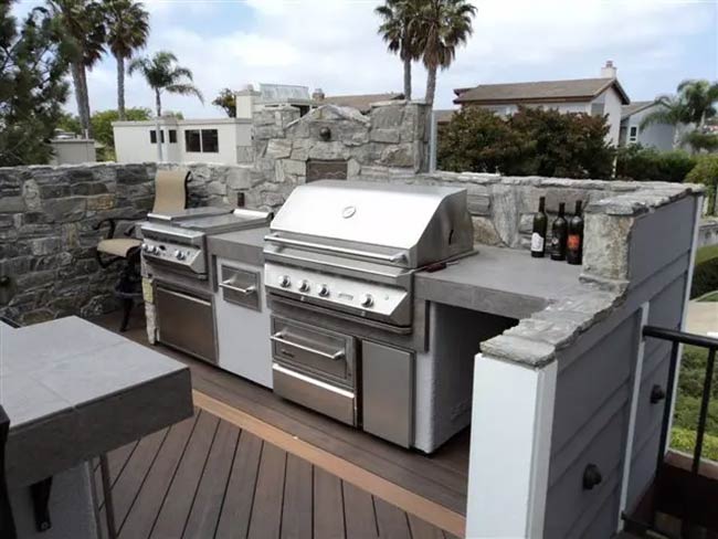 A low stone wall surrounds this rooftop grilling station to block the wind. The grey island matches the decking perfectly and holds a large stainless steel barbecue. You can see palm trees in the background, adding a tropical vibe.