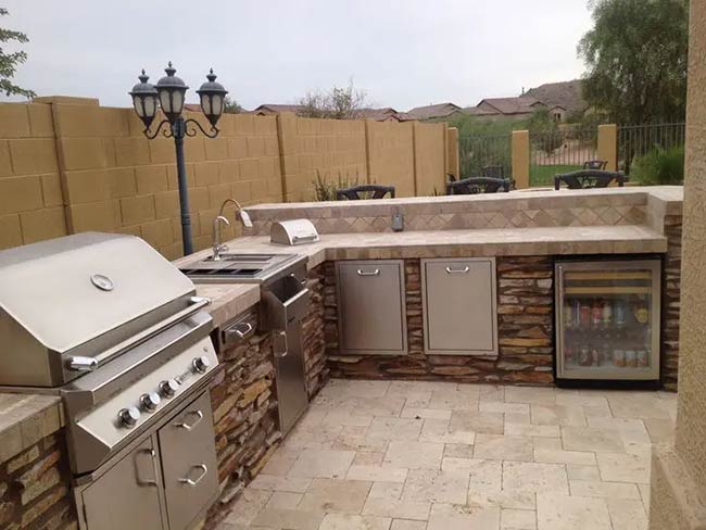 Rough stone veneer and tile countertops give this corner kitchen a very traditional feel. It has everything you need, including a sink, grill, and a glass-door fridge. The tall street lamps add a classic touch to the patio.