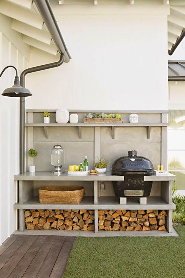 A grill sits next to the regular gas barbecue for serious cooking. The concrete countertop provides plenty of space for prepping food, while wood storage underneath keeps logs dry. It’s a lovely farmhouse-style setup on a grassy patio.