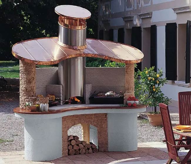 This unique curved kitchen features a tall copper chimney for the wood-fired oven. Stone columns and a split-level counter create a very traditional and sturdy look. It feels like a little castle in the garden.