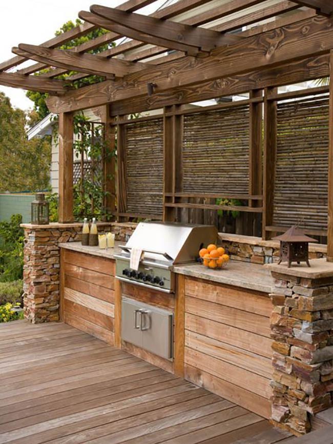 Warm wood panels and stacked stone pillars give this kitchen a natural, rustic charm. The built-in grill is surrounded by plenty of counter space for serving drinks and food. A wooden pergola overhead provides just enough shade for the chef.