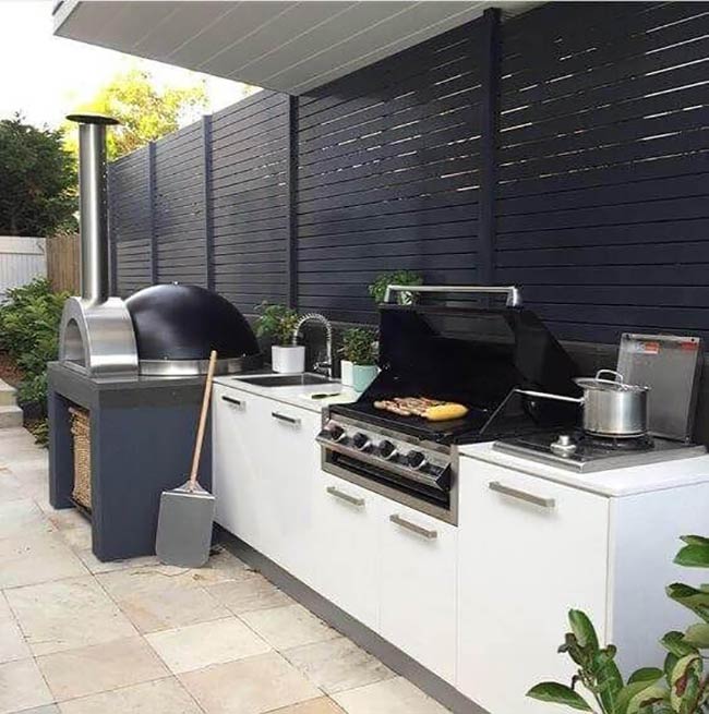 The tall silver chimney of the pizza oven stands out against the dark fence. White cabinets keep the look clean and bright on the stone patio. It is a great compact kitchen for a small backyard.