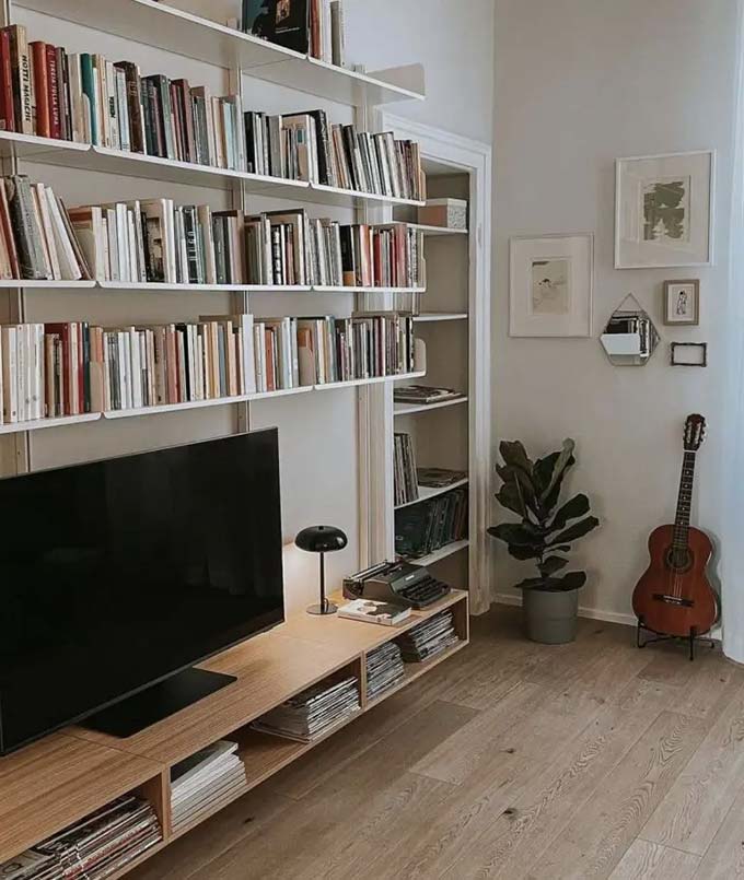 White shelves go all the way to the ceiling to hold many colorful books. The TV sits low on a long wooden bench that matches the floor. A guitar in the corner adds a fun personal touch. This setup is perfect for anyone who needs lots of storage space.