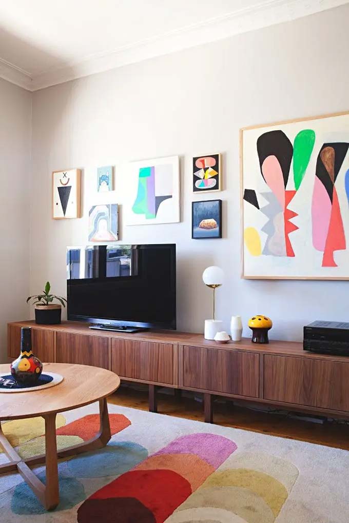 A very long wooden console stretches across the room to ground the TV area and provide storage. Bright, colorful abstract paintings hang on the white wall to make the space feel fun and energetic. This modern TV wall decor keeps things simple but very lively.