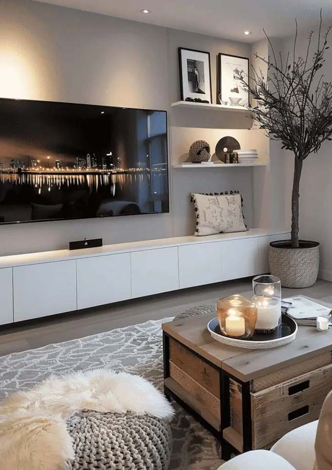A very long, white floating cabinet runs across the wall to create a clean and modern look. Simple black and white art leans on a shelf to the side of the large screen. The tall tree in the corner adds height and life to this minimalist TV wall decor.