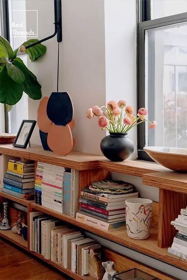 Simple plywood shelves wrap right around the corner to connect the two walls. It is a very artistic way to display colorful books and pretty vases. The design adds warm wood tones and useful under window storage to the bright room.